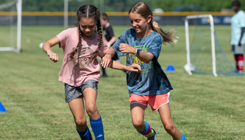 Two girls running on a soccer field, smiling and competing closely.