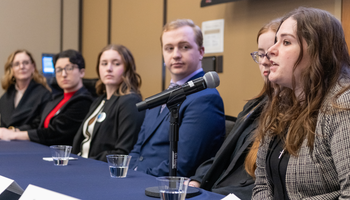 Panel of six people sitting at a table, one speaking into a microphone.