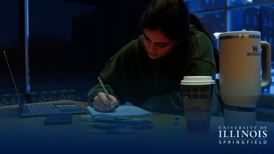 Student studying at a desk with a laptop and coffee cup, in dim lighting.