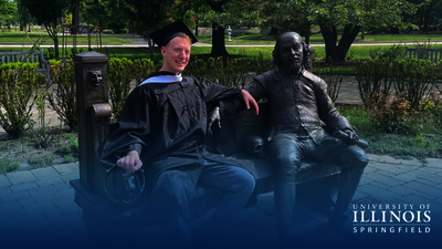 Graduate sitting next to a statue on a bench in a park setting, smiling.