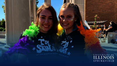 Two people smiling, wearing rainbow feather boas outdoors in sunlight. Their t-shirts say Tri Sigma.