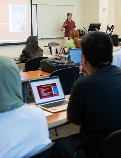 The backs of heads of students sitting in a classroom listing to a professor teach. Several laptop computers are visible.