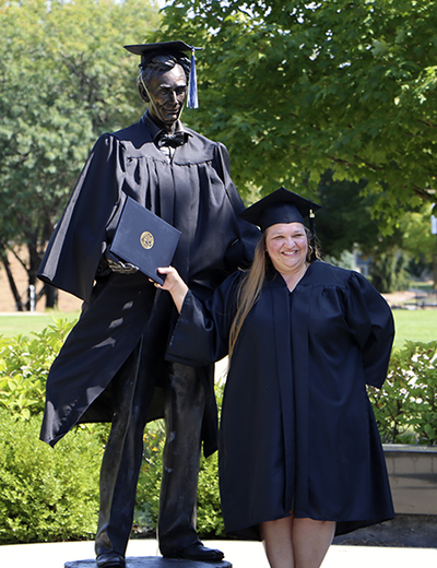 A student in cap and gown stands next to the Abraham Lincoln statue on campus (also in cap and gown).