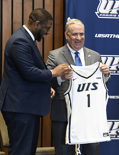 Mike Hermann holding a basketball jersey reading "UIS 1" stands next to Vice Chancellor Jamarco Clark on stage.