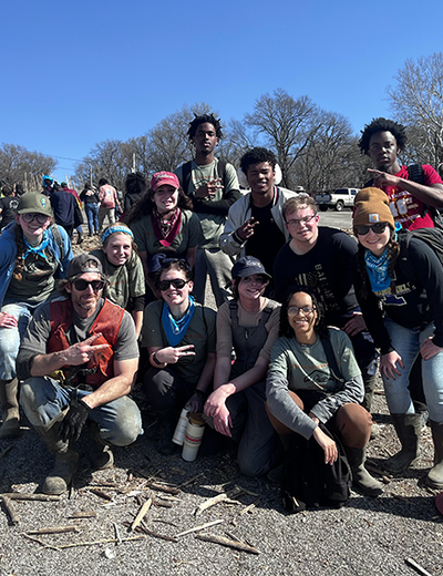 A group of volunteers poses for a photo during an outdoor cleanup event, wearing work gloves, boots, and bandanas. A damaged boat and debris are visible in the background.