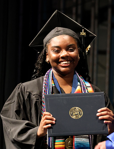 A UIS graduate smiles while holding her diploma on stage during the commencement ceremony, wearing a cap and gown.