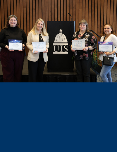 Four award recipients pose with their certificates in front of a UIS podium at a recognition event.