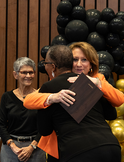 A person receives an award and a hug during the Good as Gold Volunteer Awards ceremony, with others smiling nearby.
