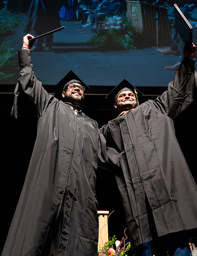Two graduates in black caps and gowns celebrate on stage by raising their diplomas triumphantly, smiling as they stand side by side. Behind them, faculty members in academic regalia sit in rows and a large screen displays a projection of the moment above them.