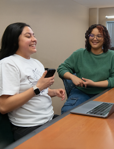 Students laugh and talk in a classroom setting with laptops on desks and a relaxed, collaborative atmosphere.