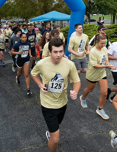 A group of runners participates in a race, starting under a blue inflatable arch. Participants wear numbered bibs and light-colored shirts.