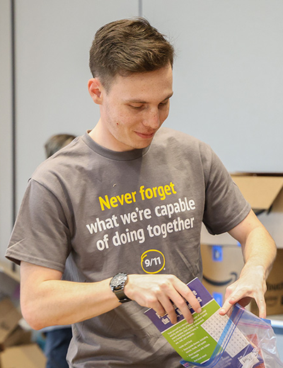 A person packing a colorful bag with supplies in a room filled with boxes. Their shirt reads, "Never forget what we're capable of doing together." 
