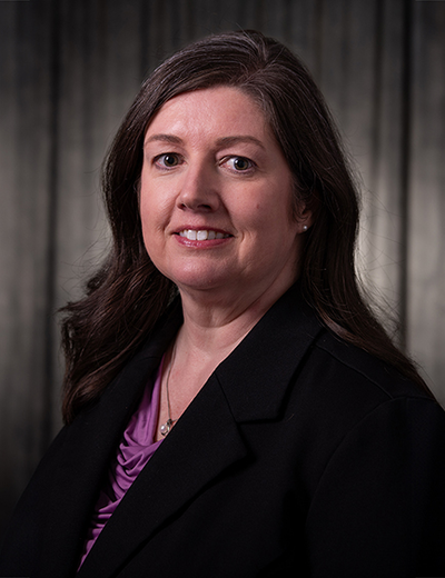 Smiling woman with dark hair in a dark room, wearing a purple shirt.