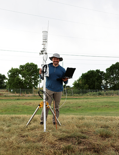 Man with tablet stands by weather station in grassy field.