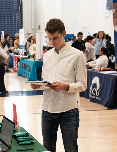 Young man reading a brochure at a busy indoor event with booths.