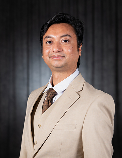 Man in a tan suit with a tie, black background, smiling gently.