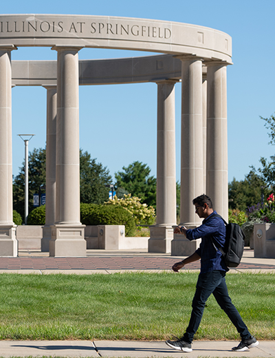 Man walks past stone pillars in park setting under clear blue sky.