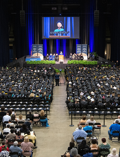 Graduation ceremony with speaker on stage, audience in caps and gowns.