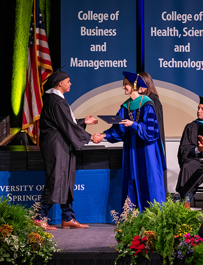 Graduate receives diploma on stage from faculty member in a blue robe.