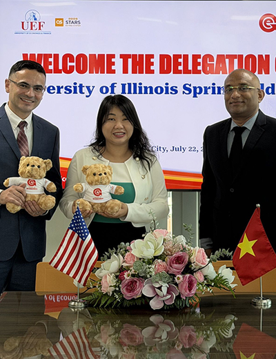 Three people smiling, holding teddy bears, with flags and flowers on a table.