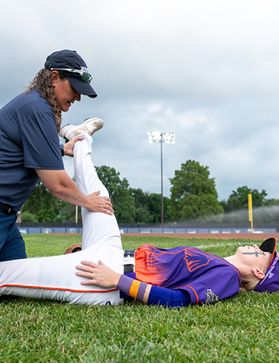Baseball coach helps player stretch leg on field, cloudy sky.