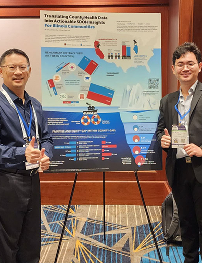 Two men smiling and giving thumbs up, standing beside a colorful conference poster on a stand.