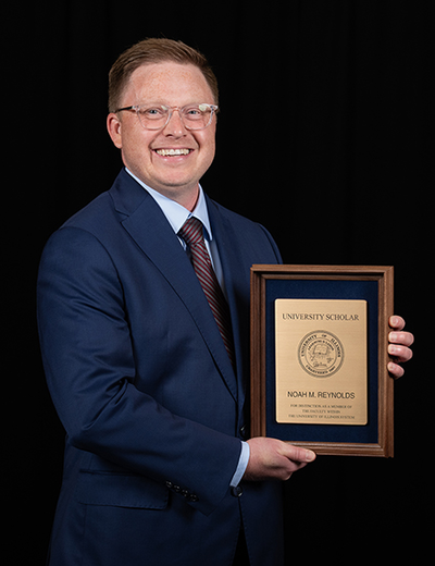 Man in suit holding a wooden plaque, smiling against a black background.
