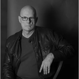 Man in a leather jacket, sitting in dim lighting, looking serious.