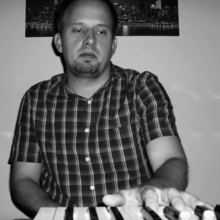 Man in plaid shirt playing keyboard in dim light, looking focused.