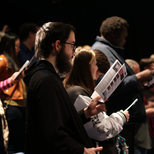 People standing and singing from sheets in a concert hall.