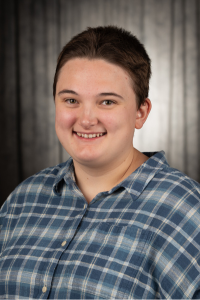 Morell Old, smiling in a blue plaid shirt against a dark background.