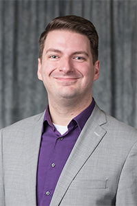 Smiling man in a gray suit and purple shirt against a dark curtain.