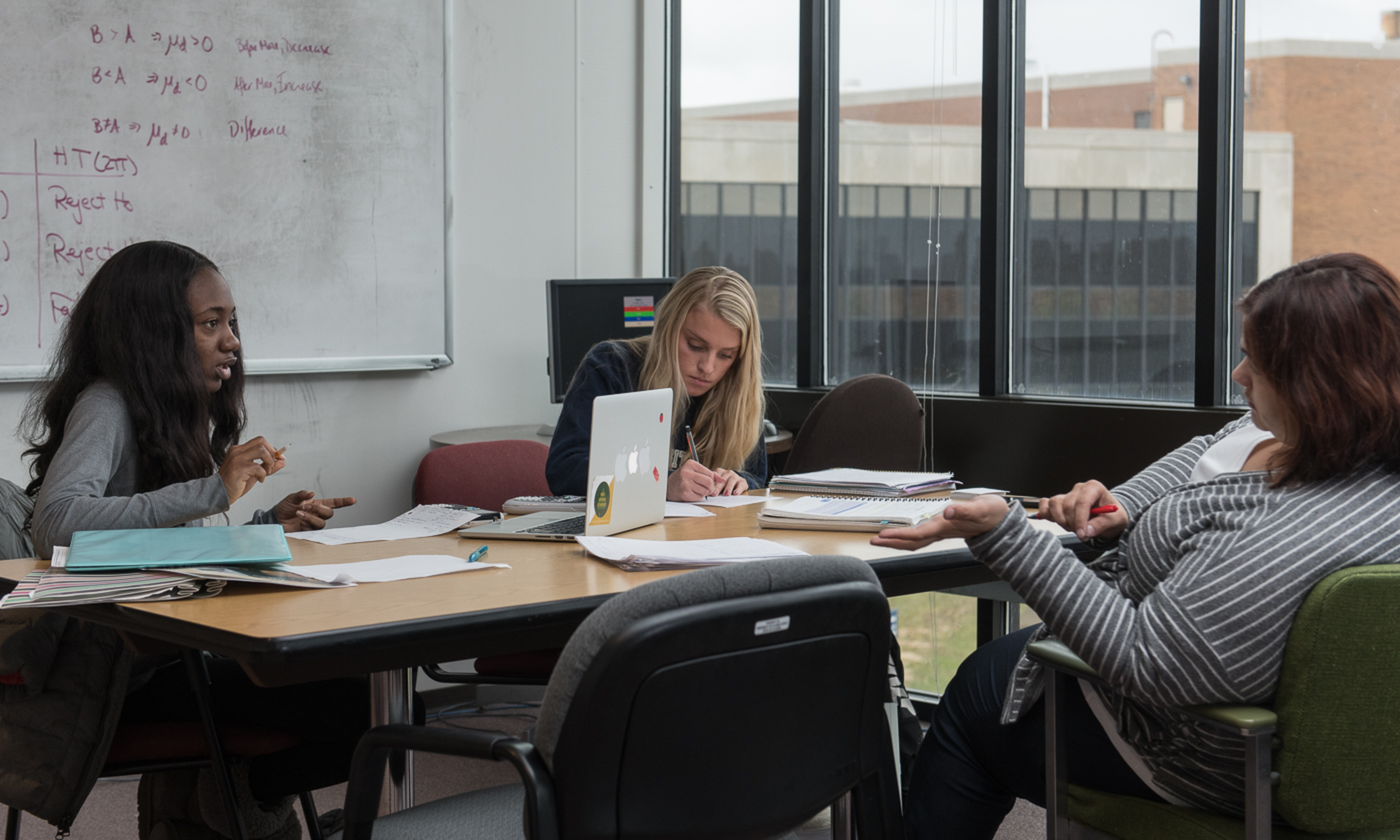 tutor and two students sit at a desk with homework spread out, and a whiteboard behind them with information written out