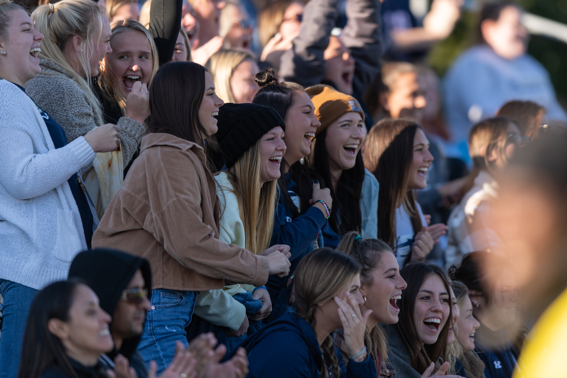 A group of students laughs and cheers for the soccer team in fall clothes and stocking caps.