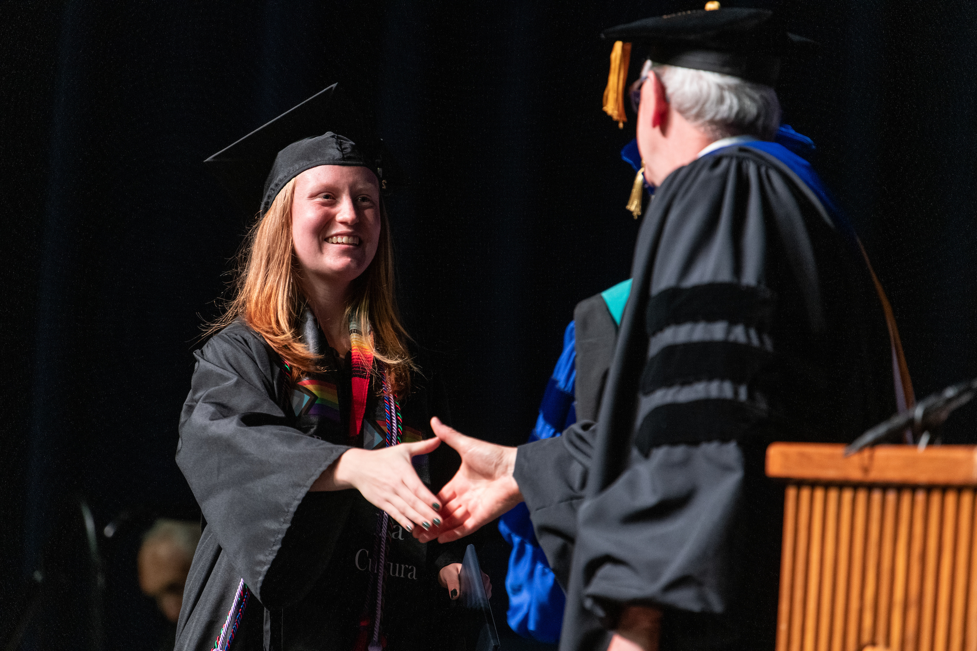 Young woman with red hair wearing a cap and mortar board shakes the hand of an administrator while on stage at Commencement.
