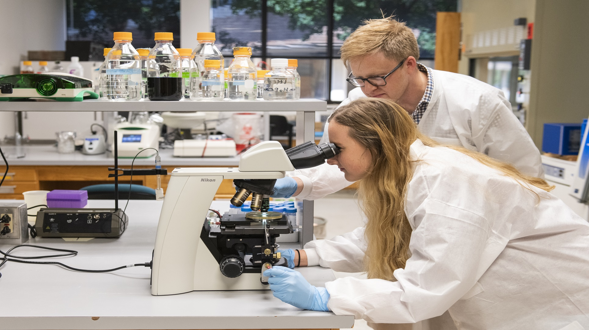Two people in lab examining things under a microscope in a lab.