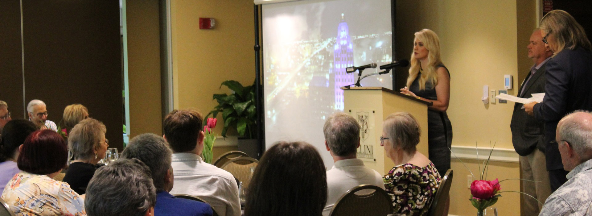 a large group of people sit at tables while a woman gives a presentation
