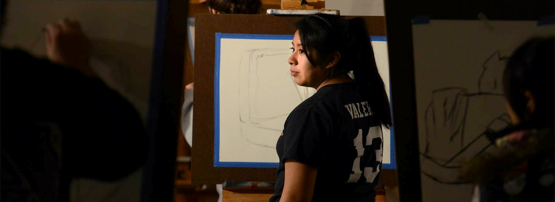 an art student looking at a still life subject while drawing it on her canvas on an easel