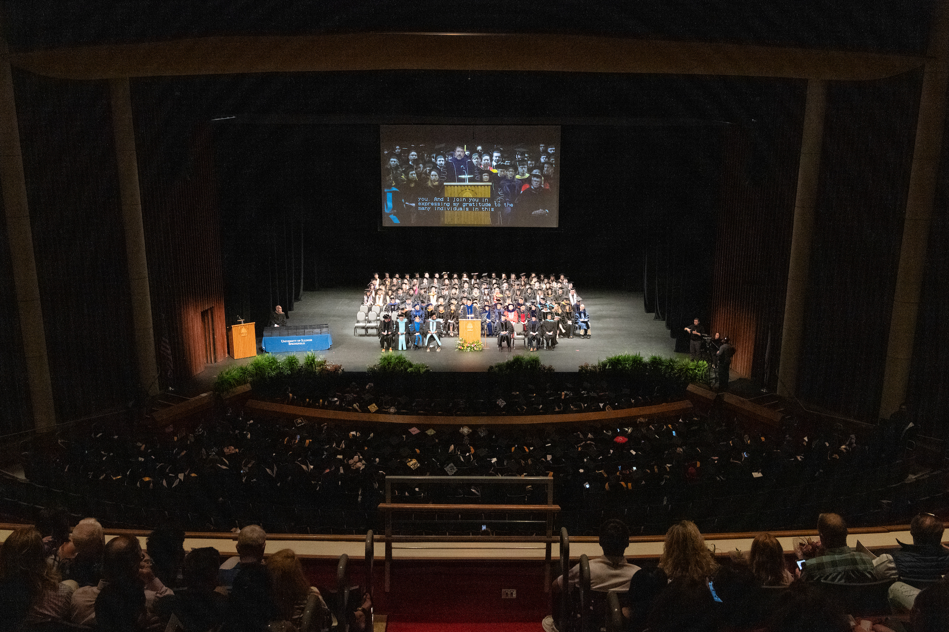 A view of the College of Health, Science and Technology graduates seated on stage in regalia on the stage of the Sangamon Auditorium with the audience in the foreground.