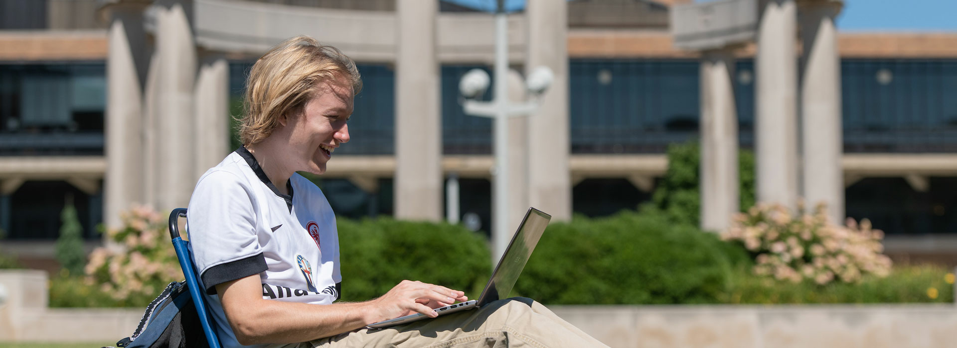 a young man sitting on the quad laughing while looking at his laptop