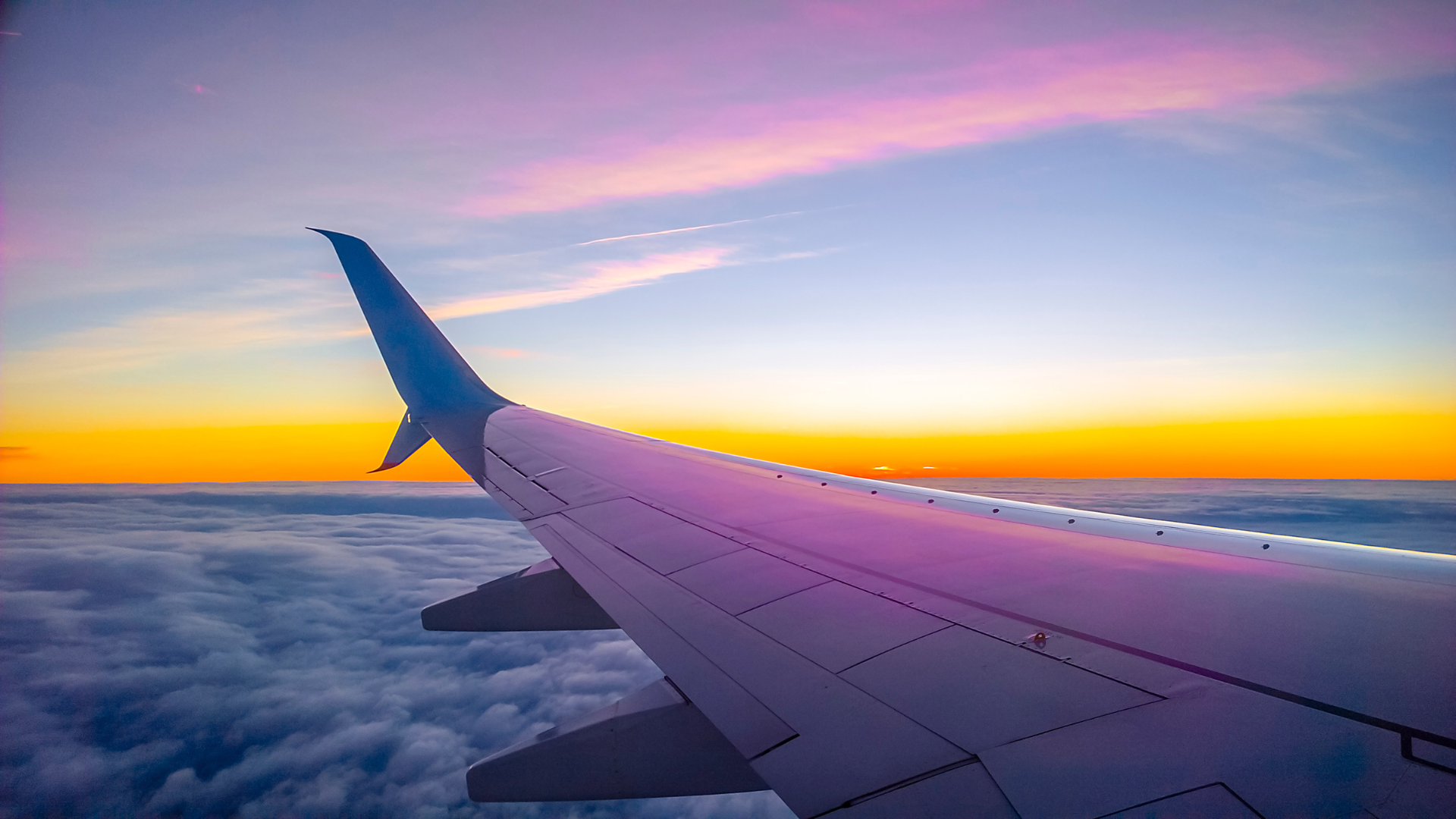 A view from a plane window while the plane is in flight. The wing is visible, there is and orange and pink sunset on the horizon with clouds underneath.