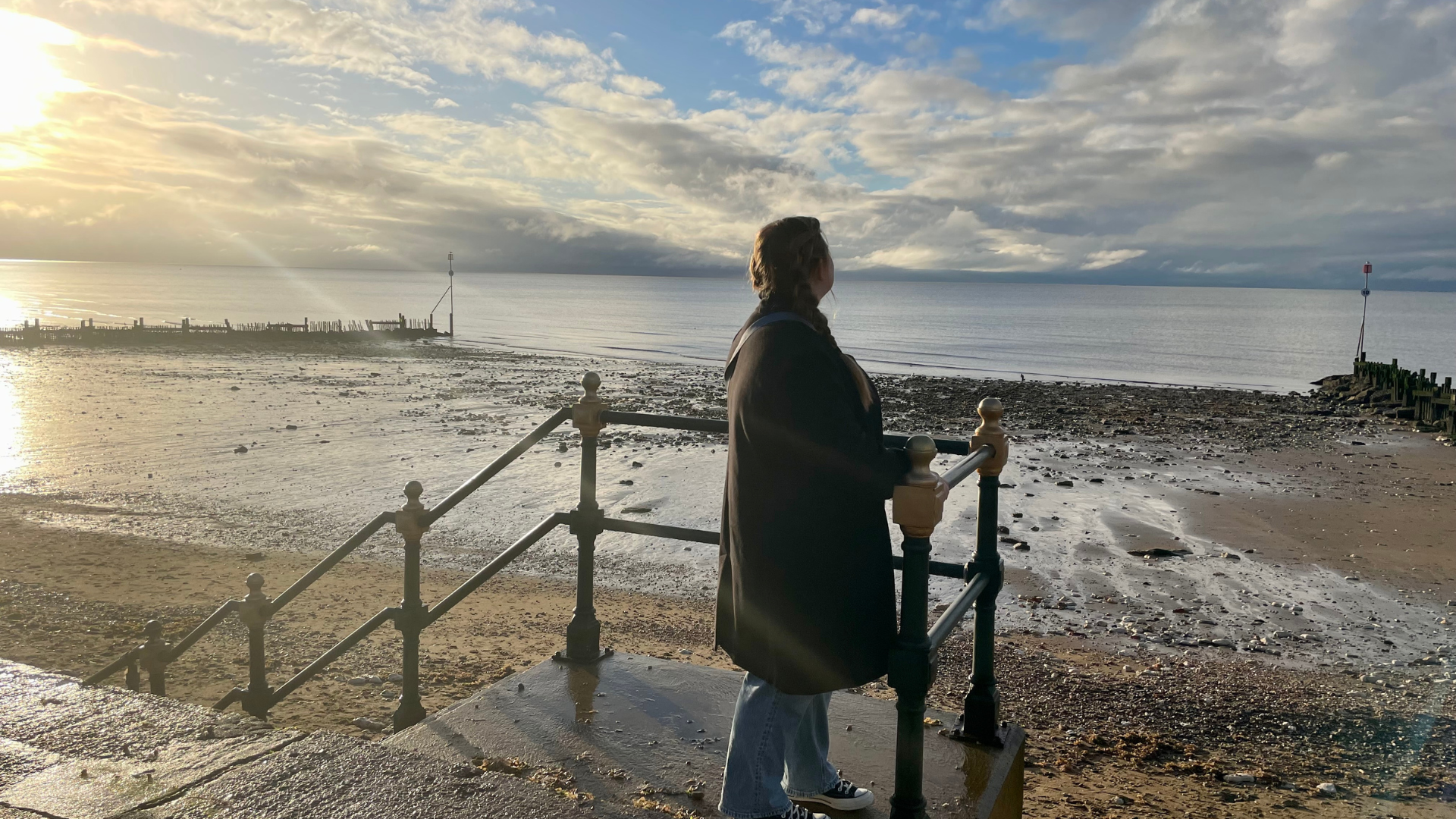 Person standing on a staircase and facing the beach below. On the left, there is a distant dock on the water and a sun beginning to set in the sky.