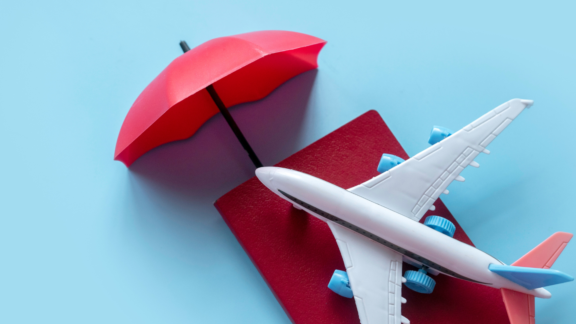 Blue background. Small red passport with a toy airplane on top of it. There is a red toy umbrella coming out of the top of the passport.