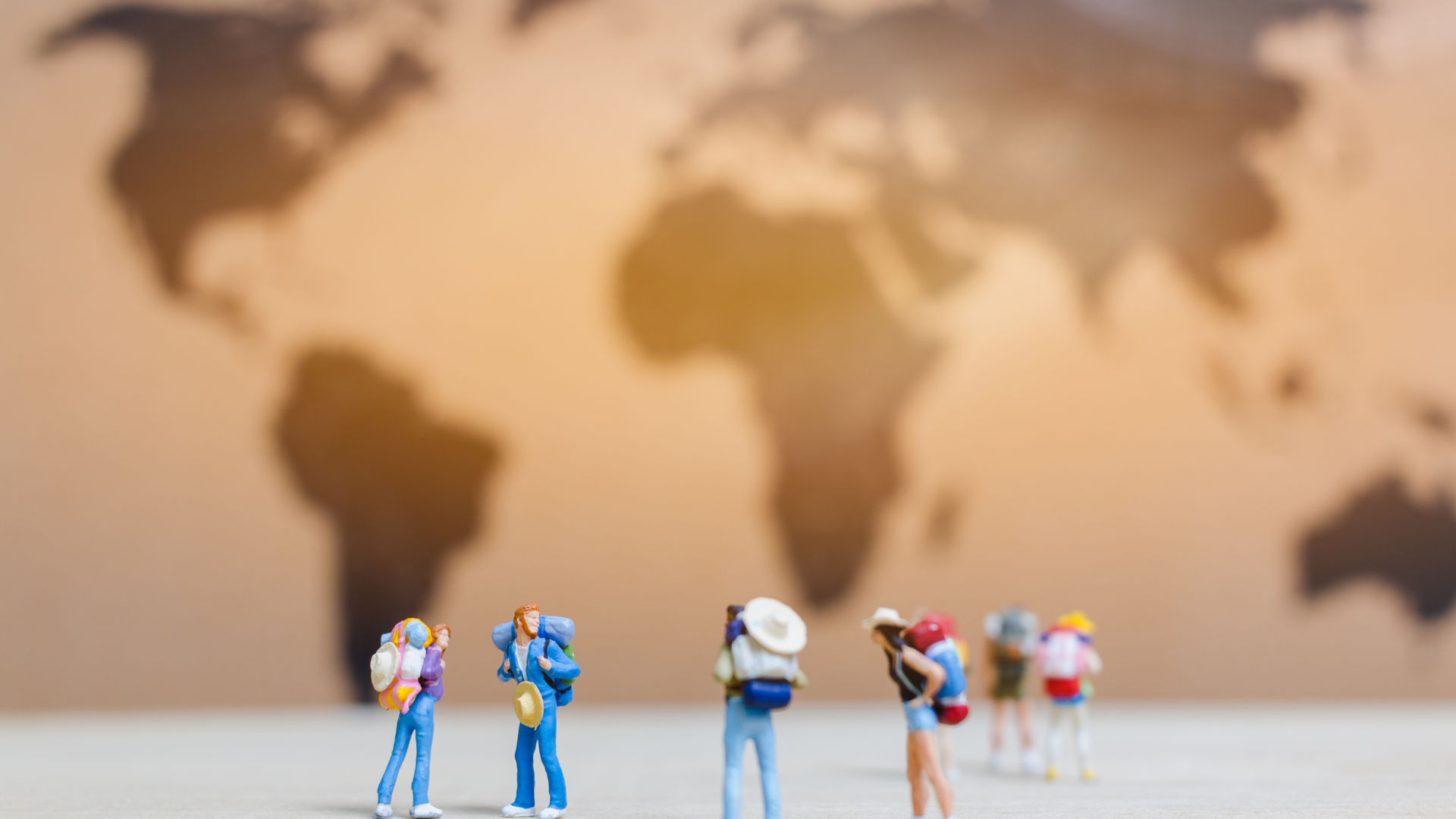 Numerous plastic figurines with travel paraphernalia on their person. The figurines are standing on a white ground and face a brown world map that is out of focus.