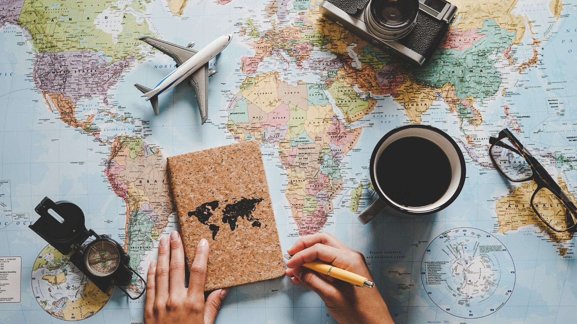 World map with a coffee cup and reading glasses resting on it. There is also travel paraphernalia scattered across the map: a compass, camera, toy airplane, and a small notebook with some continents on the cover. There is a person holding a pencil in one hand while their other hand appears to be opening the notebook.   