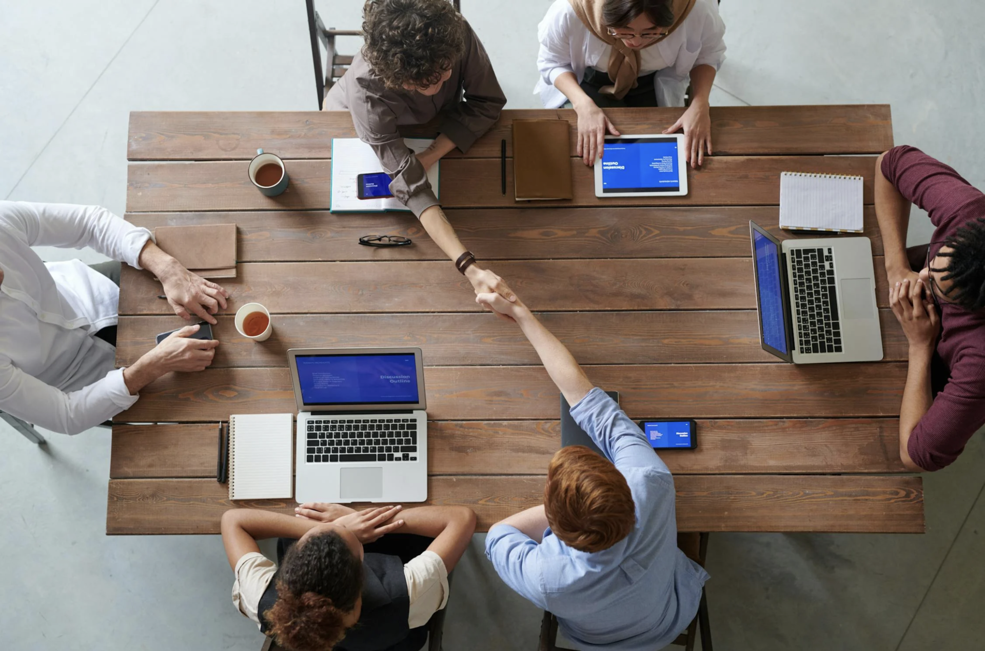 Six members sitting on a working table with 3 laptops and 2 members are giving a handshake 