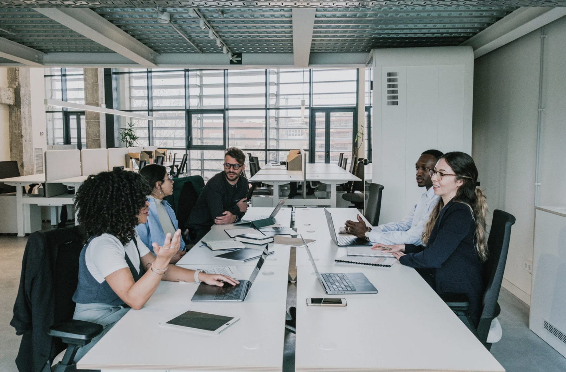 5 members sitting with laptops in front of them in an office space and discussing with each other.