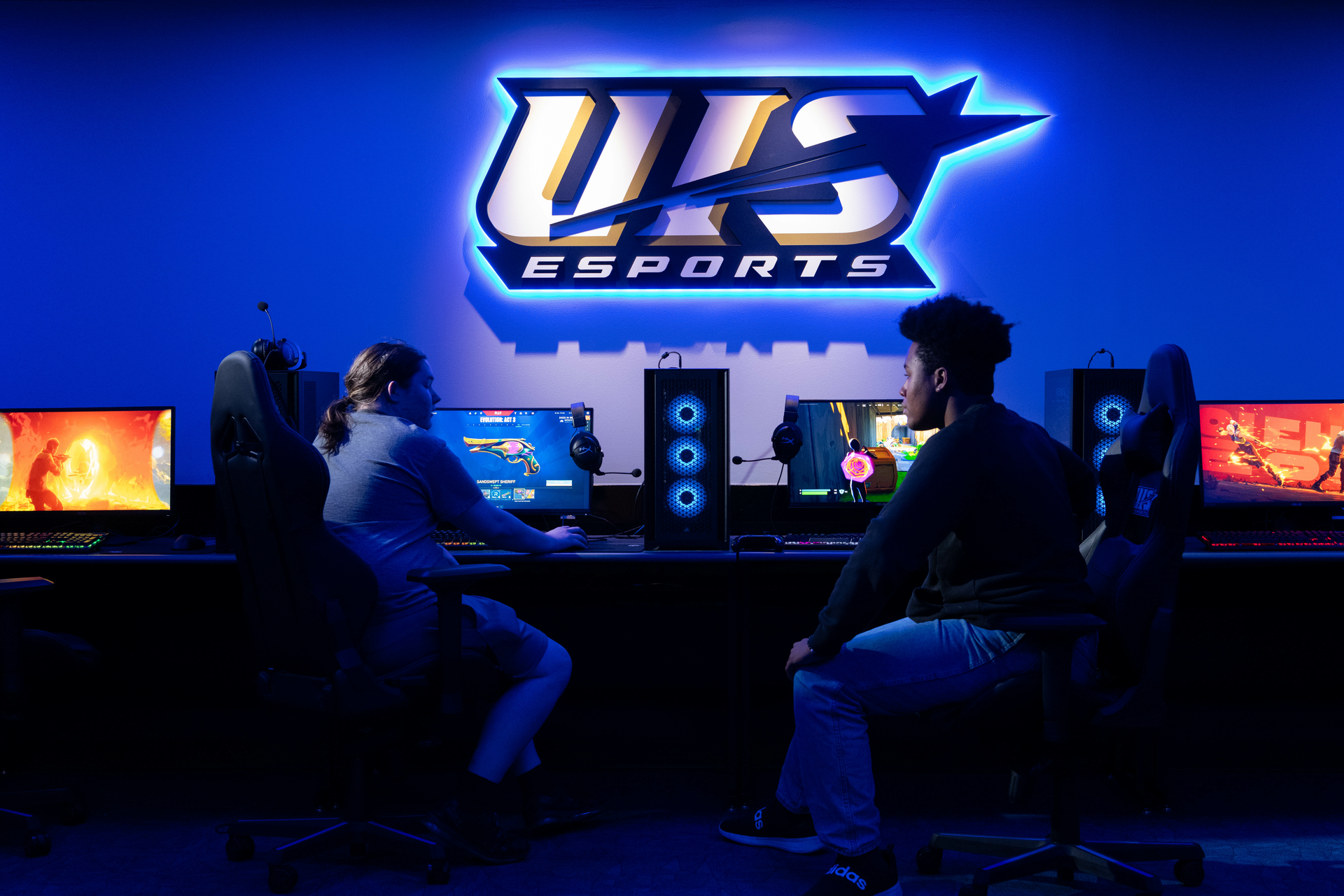 Two students sitting at computers in the Esports Arena in front of the light up UIS Esports sign. 