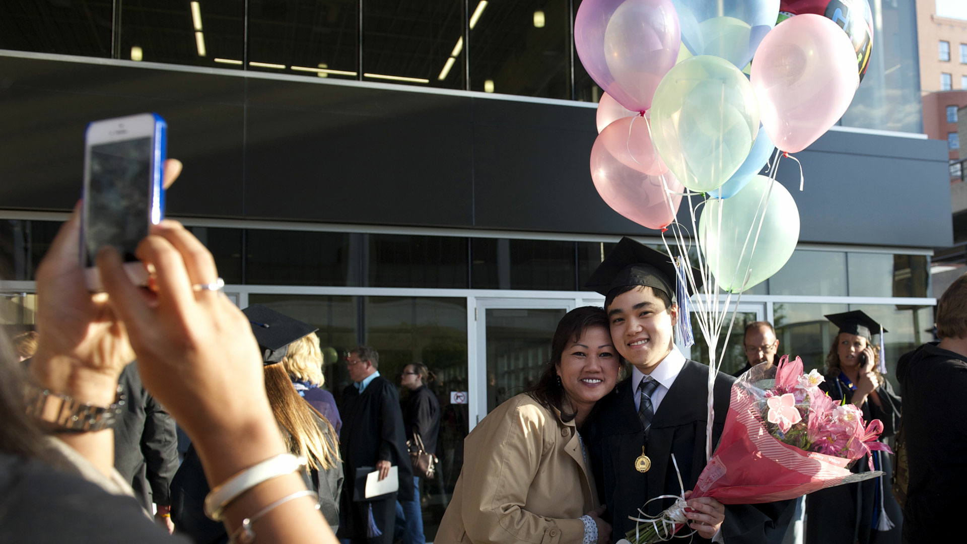 a male graduate in cap and gown holding balloons while posing with his mom
