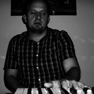 Man in plaid shirt playing keyboard in dim light, looking focused.